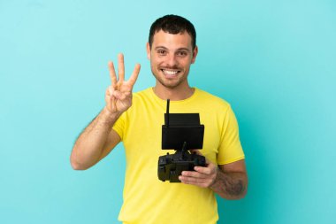 İzole edilmiş mavi arkaplan üzerinde uzaktan kumanda tutan Brezilyalı adam mutlu ve parmaklarıyla üçe kadar sayıyor.