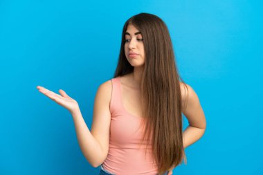 Mavi arka planda izole edilmiş genç beyaz bir kadın. Kopyalama alanını şüphe ile tutuyor.