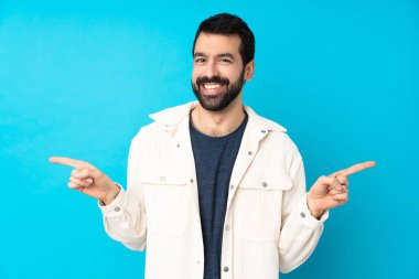 Mavi arka planda beyaz kadife ceketli genç yakışıklı adam parmak uçlarını işaret ediyor ve mutlu.