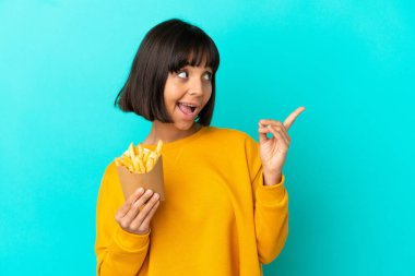 İzole edilmiş mavi arka planda patates kızartması tutan genç esmer kadın çözümü bulmak için parmağını kaldırıyor.