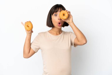 Genç, melez, hamile bir kadın, beyaz arka planda gözünün içinde donut tutarak izole edilmiş.