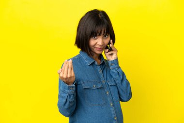 Sarı arka planda izole edilmiş cep telefonu kullanan hamile bir kadın eliyle gelmesi için davet ediyor. Gelmene sevindim.