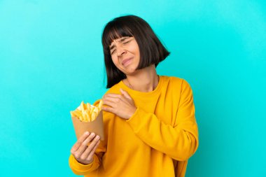 Genç esmer kadın, izole edilmiş mavi arka planda patates kızartması tutuyor. Çabaladığı için omuz ağrısı çekiyor.