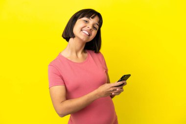 Genç melez hamile kadın sarı arka planda izole edilmiş cep telefonuyla mesaj ya da e-posta gönderiyor.