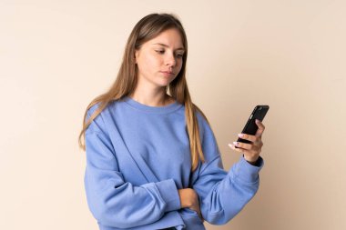 Bej arka planda izole edilmiş, üzgün ifadeyle cep telefonu kullanan Litvanyalı genç bir kadın.