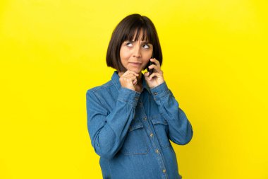 Sarı arka planda izole edilmiş cep telefonu kullanan hamile kadının şüpheleri ve düşünceleri var.