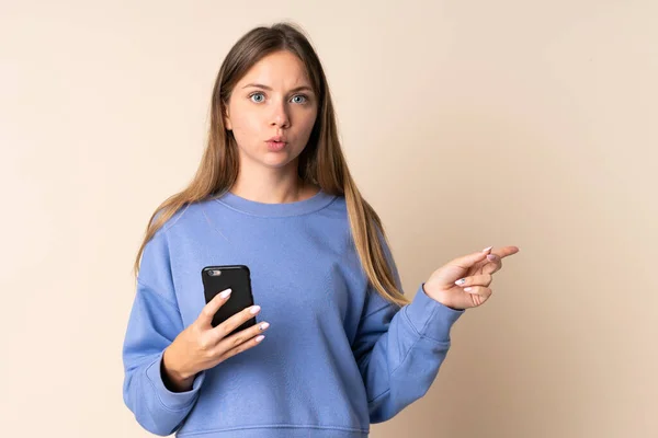 Молодая Литовская Женщина Использующая Мобильный Телефон Изолирован Бежевом Фоне Удивлен — стоковое фото