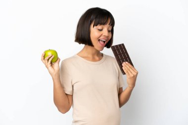 Genç, melez, hamile bir kadın beyaz arka planda izole edilmiş. Bir elinde çikolata tableti, diğer elinde elma var.