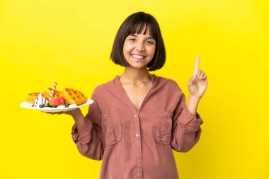 Sarı arka planda izole edilmiş waffle 'lar tutan hamile bir kadın parmağını yana uzattı ve bir ürün sundu.