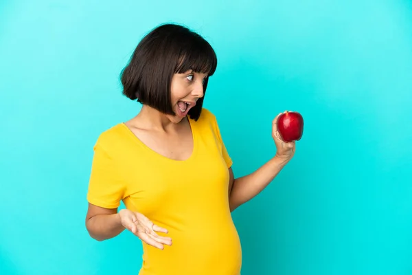妊娠中の女性側を見ながら驚きの表情で青の背景に隔離されたリンゴを保持 — ストック写真