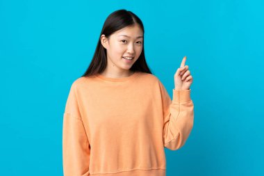 Genç Çinli kız, izole edilmiş mavi arka planda en iyinin işareti olarak parmağını kaldırıyor.