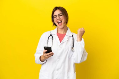 Doktor kadın sarı arka planda izole edilmiş ve telefon zafer pozisyonunda.