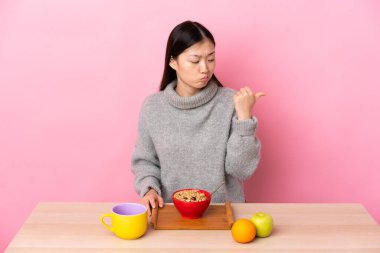 Genç Çinli kız mutsuz bir masada kahvaltı yapıyor ve yan tarafı işaret ediyor.