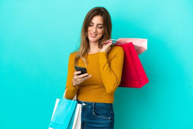 Beyaz bir kadın mavi arka planda izole edilmiş. Elinde alışveriş poşetleri var ve bir arkadaşına cep telefonuyla mesaj yazıyor.
