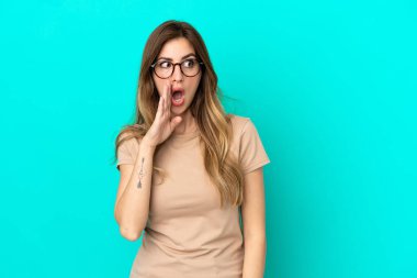Mavi arka planda izole edilmiş genç beyaz kadın yan tarafa bakarken sürpriz bir jest yaparak bir şeyler fısıldıyor.