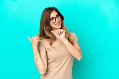 Mavi arka planda izole edilmiş genç beyaz kadın bir ürünü sunmak için kenarı işaret ediyor.