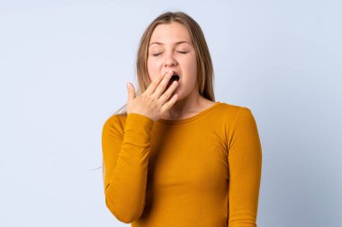 Genç Ukraynalı kız mavi arka planda izole edilmiş esniyor ve ağzını eliyle açık tutuyor.