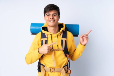 İzole edilmiş mavi arka plan üzerinde büyük bir sırt çantası olan genç dağcı, parmağını yana doğru gösteriyor.