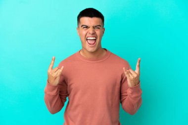 Soyutlanmış mavi arka planda genç yakışıklı adam boynuz hareketi yapıyor.