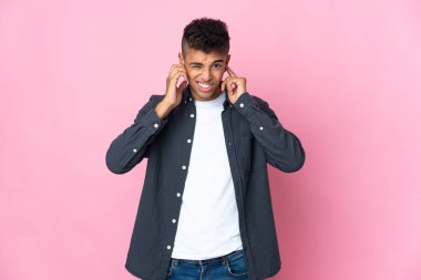 Pembe arka planda izole edilmiş genç Brezilyalı adam hayal kırıklığına uğramış ve kulaklarını kapatmış.