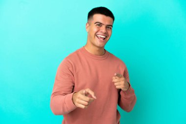 Soyutlanmış mavi arka planda genç yakışıklı adam şaşırmış ve ön tarafı işaret ediyor.