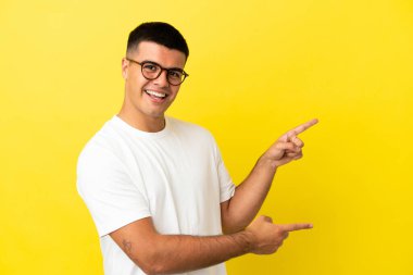 İzole edilmiş sarı arka planda genç yakışıklı bir adam parmağını yana uzatıyor ve bir ürün sunuyor.