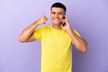 Genç yakışıklı adam, izole edilmiş mor arka plan üzerinden cep telefonu kullanıyor ve telefon jesti yapıyor. Bana geri dönüş işareti ver.