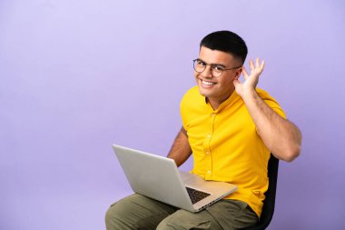 Genç adam dizüstü bilgisayarlı bir sandalyede oturuyor ve elini kulağına koyup bir şeyler dinliyor.