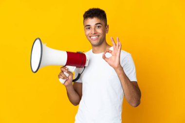 Sarı arka planda izole edilmiş genç Brezilyalı bir adam elinde bir megafonla tamam işaretini parmaklarıyla gösteriyor.