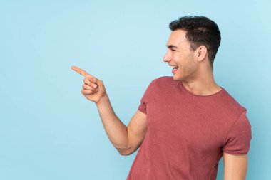 Mavi arka planda izole edilmiş beyaz tenli yakışıklı bir adam, parmağını yana uzattı ve bir ürün sundu.