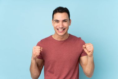 Beyaz tenli yakışıklı adam mavi arka planda izole edilmiş ve galibiyeti kutluyor.