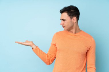 Mavi arka planda izole edilmiş beyaz, yakışıklı bir genç. Kopyalama alanını şüphe ile tutuyor.