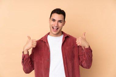 Beyaz tenli yakışıklı adam bej arka planda izole edilmiş. Baş parmakları havada ve gülümsüyor.