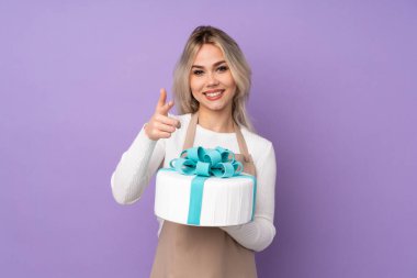 Genç pasta şefi mor arka planda büyük bir pasta tutuyor. Parmağını sana gösteriyor.