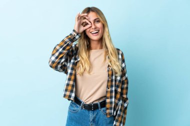 Mavi arka planda izole edilmiş genç sarışın Uruguaylı parmaklarıyla tamam işareti gösteriyor