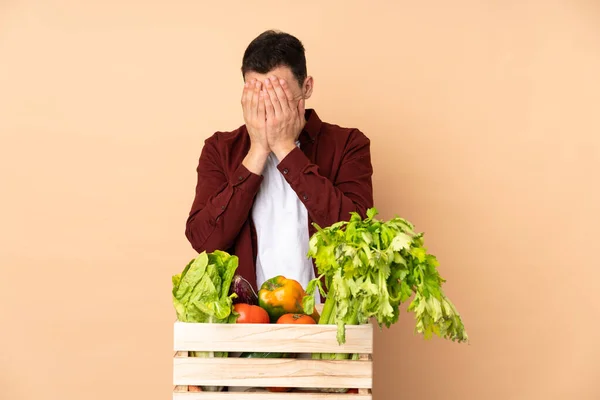 Farmer with freshly picked vegetables in a box isolated on beige background with tired and sick expression