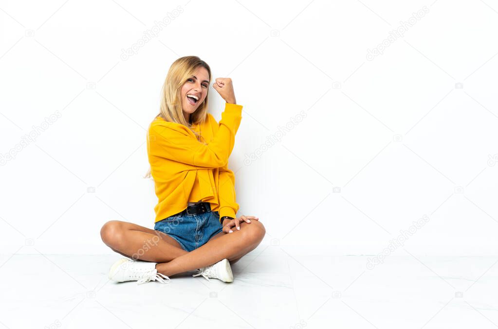 Young blonde Uruguayan woman sitting on the floor isolated on white background doing strong gesture