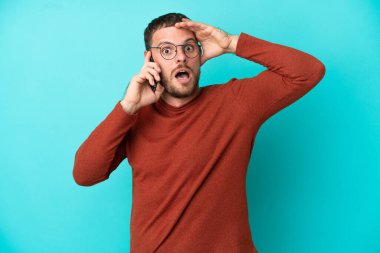 Mavi arka planda izole edilmiş cep telefonu kullanan genç Brezilyalı adam yan tarafa bakarken sürpriz jest yapıyor.