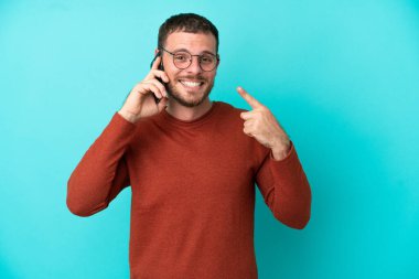Mavi arka planda izole edilmiş cep telefonu kullanan genç Brezilyalı adam işaret veriyor.