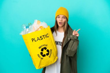 Elinde plastik şişelerle dolu bir çanta tutan küçük bir kız izole edilmiş mavi arka planda geri dönüşüm yapmak için parmağını kaldıran bir fikir düşünüyor.