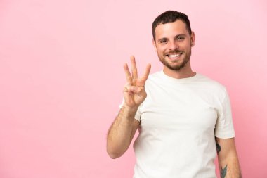 Pembe arka planda izole edilmiş genç Brezilyalı mutlu ve parmaklarıyla üçe kadar sayıyor.