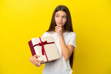 Küçük kız sarı arka planda bir hediye tutuyor ve yukarı bakıyor.