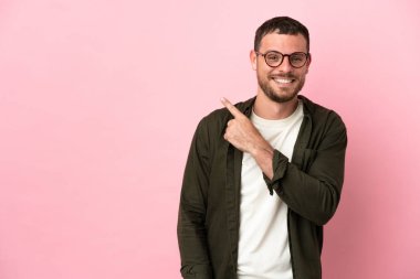 Pembe arka planda izole edilmiş genç Brezilyalı bir adam bir ürün sunmak için kenarı işaret ediyor.