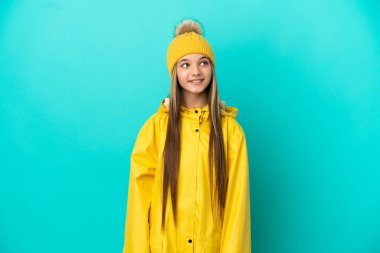 İzole edilmiş mavi arka planda, yağmura dayanıklı bir ceket giyen küçük kız yukarı bakarken bir fikir düşünüyor.