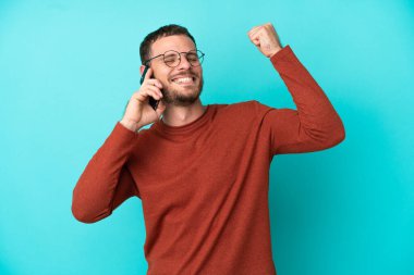 Mavi arka planda izole edilmiş cep telefonu kullanan genç Brezilyalı bir adam zaferi kutluyor.