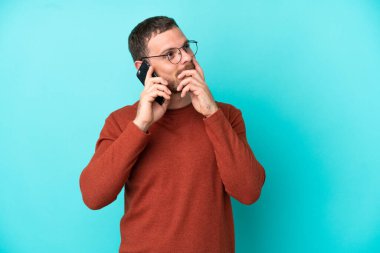 Mavi arka planda izole edilmiş cep telefonu kullanan genç Brezilyalı adam gülümserken yukarı bakıyor