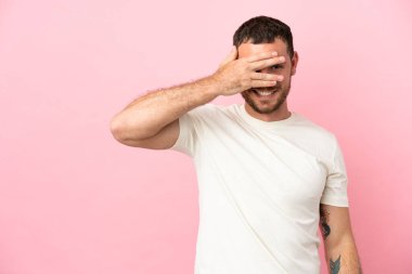 Pembe arka planda izole edilmiş genç Brezilyalı adam gözlerini elleriyle kapatıp gülümsüyor.