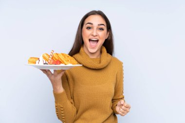 Mavi arka planda, şaşırmış ve şok olmuş bir yüz ifadesiyle waffle tutan beyaz bir kadın.