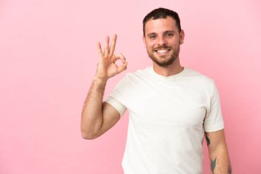 Pembe arka planda izole edilmiş genç Brezilyalı adam parmaklarıyla tamam işareti gösteriyor.