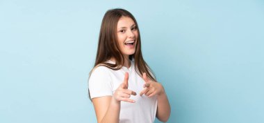 Genç beyaz kız, mavi arka planda izole edilmiş. Önü işaret ediyor ve gülümsüyor.